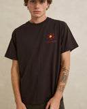 A man wearing a dark brown short-sleeve t-shirt with a small red and yellow graphic on the left chest, photographed against a neutral beige background. Rhythm Mens Cactus SS T-Shirt - Vintage Black