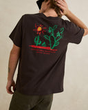 A man wearing a dark brown t-shirt with a colorful graphic of a cactus, sun, and the text 'GOT THE WILL, WHO HAS FAITH?' on the back, paired with olive green pants against a neutral beige background. Rhythm Mens Cactus SS T-Shirt - Vintage Black