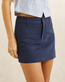 A close-up of a model wearing a navy blue mini skirt with a button closure and belt loops, paired with a blue and white striped shirt against a light beige background. Rhythm Canyon Mini Skirt