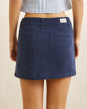 A model wearing a navy blue denim mini skirt with two front patch pockets and a white brand label on the back waistband, paired with a blue and white striped crop top against a neutral beige background. Rhythm Canyon Mini Skirt