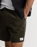 Close-up of a person wearing dark olive corduroy shorts with a white brand patch on the hem, styled with a white t-shirt and a dark overshirt against a light gray background. Rhythm Mens Classic Chord Jam - Olive