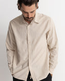 A man wearing a light beige linen button-down shirt, looking down with his hands in his pockets, against a plain light gray background. Rhythm Men's Classic Linen LS Shirt – Sand