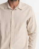 A close-up of a man wearing a light beige linen button-down shirt, showing the collar, front buttons, and textured fabric against a plain white background. Rhythm Men's Classic Linen LS Shirt – Sand