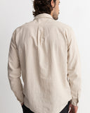 A man viewed from the back wearing a light beige linen button-down shirt, paired with black trousers, against a plain white studio background. Rhythm Men's Classic Linen LS Shirt – Sand