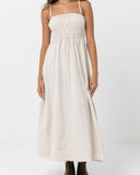 A woman modeling a light beige linen maxi dress with a smocked bodice and thin spaghetti straps, shown against a plain white studio background. Rhythm Classic Shirred Midi Dress - Oat