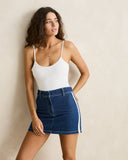 A woman modeling a white camisole top and a dark blue denim mini skirt with white side stripes, standing against a plain beige wall.