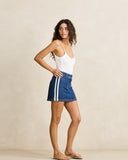 A woman modeling a white ribbed tank top and a blue denim mini skirt with white side stripes, standing against a warm beige textured studio wall.