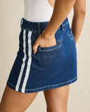 A close-up of a model wearing a blue denim mini skirt with two vertical white stripes down the side and a front pocket, paired with a white top against a light beige background.