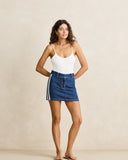 A woman modeling a white spaghetti-strap bodysuit and a blue denim mini skirt with white side stripes, standing against a neutral beige studio backdrop.