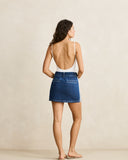 A woman shown from the back wearing a blue denim mini skirt and a white open-back camisole, standing against a plain beige studio wall.