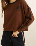 A person wearing a cropped brown sweatshirt with black trim at the collar and double black stripes down the sleeves, paired with dark shorts against a plain light background. Front view of the Rhythm Contrast Slouchy Crew Neck Fleece in Chocolate brown showing the oversized fit and chest embroidery.