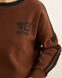 A close-up of a brown sweatshirt with a dark navy graphic on the chest featuring flower motifs and the word 'RHYTHM', along with dark navy stripes down the sleeves.