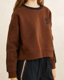 A cropped brown sweatshirt with a black crew neck and black side stripes on the sleeves, featuring a black graphic logo on the left chest, worn by a model.