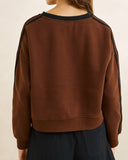 A person shown from the back wearing a brown crewneck sweatshirt with black shoulder and sleeve stripes, paired with dark pants, against a plain light beige background. Back view of the Rhythm Contrast Slouchy Crew Neck Fleece in Chocolate brown highlighting the clean drop-shoulder seams and relaxed drape.