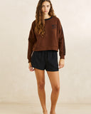 A woman modeling a brown crewneck sweatshirt with black sleeve stripes and a small black chest graphic, paired with black shorts, against a neutral beige studio backdrop.