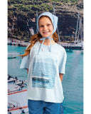 Young girl wearing a white shirt with a boat graphic and a blue bonnet, standing in front of a scenic background with boats and cliffs.