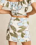 A woman wearing a matching two-piece set with a ruffled off-the-shoulder crop top and a high-waisted mini skirt, both featuring a floral print in light blue, olive green, and taupe on an off-white background. Back view of the blue floral Rhythm Gigi Skirt highlighting the straight hemline drape and clean rear profile.