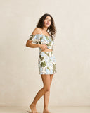 A woman modeling a matching off-the-shoulder crop top and high-waisted mini skirt set with a light blue and green tropical floral print on a cream background, set against a neutral beige studio backdrop.