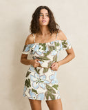 A woman modeling an off-the-shoulder floral mini dress with a cream base and light blue, green, and gray botanical print against a plain beige studio background.