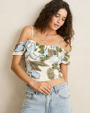 A woman wearing a cream off-the-shoulder crop top with a green and blue floral print, paired with light-wash blue jeans, posing against a neutral beige wall. Front view of the blue floral Rhythm Gigi Top showing the square neckline and relaxed tank silhouette.