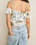A woman shown from the back wearing a cream off-the-shoulder crop top with a blue, green, and brown floral pattern, paired with light-wash denim jeans. Back view of the blue floral Rhythm Gigi Top highlighting the straight back neckline and clean drop drape.