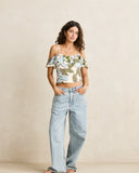 A woman modeling a cream cold-shoulder crop top with a blue and green floral print, paired with light-wash wide-leg jeans, against a neutral beige studio backdrop.
