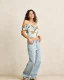 A woman modeling a light blue floral off-the-shoulder crop top with ruffled sleeves and light-wash wide-leg jeans, standing against a plain beige studio backdrop.
