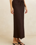 A woman wearing a dark brown textured midi skirt with a tie waist, paired with black flip-flop sandals, standing against a neutral beige wall. Rhythm Indie Maxi Skirt - Chocolate