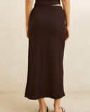 A model shown from the back wearing a dark brown ribbed knit midi skirt with a side cutout, paired with black flat sandals against a neutral beige background. Rhythm Indie Maxi Skirt - Chocolate