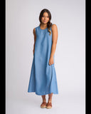A woman modeling a sleeveless, midi-length blue dress with a relaxed, A-line silhouette, paired with brown leather slide sandals, against a light gray studio background.