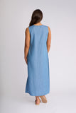 A woman viewed from behind wearing a sleeveless light blue maxi dress and beige flat sandals, standing against a plain light gray studio background.