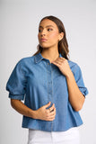 A woman wearing a cropped blue denim shirt with puff sleeves and white pants, posing against a light gray studio background.