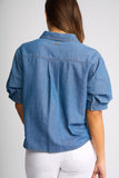 A woman viewed from the back wearing a blue denim short-sleeve button-up shirt with rolled cuffs and white pants, against a light gray studio background.