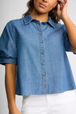 A woman wearing a cropped blue denim shirt with short puff sleeves, a pointed collar, and a button-front closure, paired with white pants against a light gray studio background.