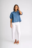 A woman modeling a cropped blue denim button-up shirt with rolled sleeves, paired with white cropped pants and tan sandals, against a light gray studio background.
