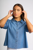 A woman wearing a blue denim short-sleeve button-up shirt, posing against a light gray studio background.