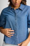 A close-up of a woman wearing a blue denim button-up shirt with short sleeves, a pointed collar, and visible contrast stitching, paired with white bottoms against a light gray studio background.