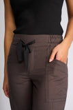 Close-up of a person wearing dark brown drawstring pants with side pockets and a black ribbed top against a light gray background. Detailed close-up macro view of the premium stretch fabrication texture, rich chocolate brown weave, and high-quality stitching on the Knewe Label pants.