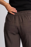 Close-up of a person wearing dark brown wide-leg trousers with an elastic waistband and large patch pockets, paired with a black ribbed top against a light gray background. Detailed close-up macro view of the premium stretch fabrication texture, rich chocolate brown weave, and high-quality stitching on the Knewe Label pants.