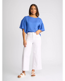 A woman modeling a bright blue short-sleeve top with voluminous flutter sleeves, paired with white wide-leg cropped pants and beige slide sandals, against a light gray studio background. Detailed close-up macro view of the premium stretch white denim fabric texture, hardware, and stitching on the Knewe Label jeans.