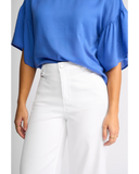 Detailed close-up macro view of the premium stretch white denim fabric texture, hardware, and stitching on the Knewe Label jeans. A person wearing a bright blue short-sleeve blouse with ruffled sleeves and white high-waisted pants, shown from the shoulders to the upper thighs against a light gray studio background.