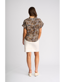A woman is shown from behind wearing a short-sleeved blouse with a brown, beige, and blue abstract pattern, paired with a cream-colored mini skirt and cream wedge sandals, set against a plain light gray studio background. Back view of the Knewe Label Addison Top in Zulu print, displaying the smooth fabric drape and pattern continuation across the shoulders.