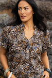 Detailed close-up macro view of the premium Zulu abstract fabric print texture and high-quality stitching on the Knewe Label blouse.  A woman wearing a short-sleeve button-front blouse with an allover leopard print pattern in brown, black, beige, and blue, photographed outdoors against a blurred rocky background.