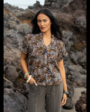 A woman wearing a short-sleeve button-up blouse with a brown, black, and white leopard print, paired with gray pants, standing on a rocky beach. Front view of the Knewe Label Addison Top in Zulu abstract print, showcasing the elegant neckline and relaxed fit layout.