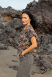 A woman stands on a rocky beach wearing a short-sleeve leopard-print blouse and gray pants, looking back over her shoulder. Side profile angle of the Zulu print Knewe Label Addison Top, highlighting the comfortable sleeve profile and fluid silhouette.