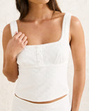 A close-up of a woman wearing a white eyelet lace cropped tank top with a square neckline and wide straps, paired with a matching white eyelet skirt against a textured white wall. Front view of the white Rhythm Lia Broderie Top showing the square neckline, eyelet detailing, and elasticated puff sleeves.