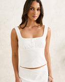 A woman wearing a white textured sleeveless crop top with a square neckline and button detail, paired with a matching white skirt, standing against a textured off-white wall. Rhythm Lia Broderie Top - White
