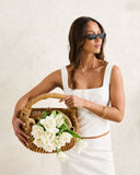A woman wearing a white textured two-piece set and black sunglasses holds a wicker basket filled with white roses against a textured off-white wall. Rhythm Lia Broderie Top - White