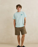 A man modeling a light blue t-shirt with a small teal graphic on the left chest, paired with brown shorts, standing against a textured beige wall. Rhythm Men's Monterey Short - Fatigue