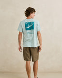 A man wearing a light blue t-shirt with a teal graphic print on the back and brown shorts, standing against a textured beige wall. Rhythm Men's Monterey Short - Fatigue